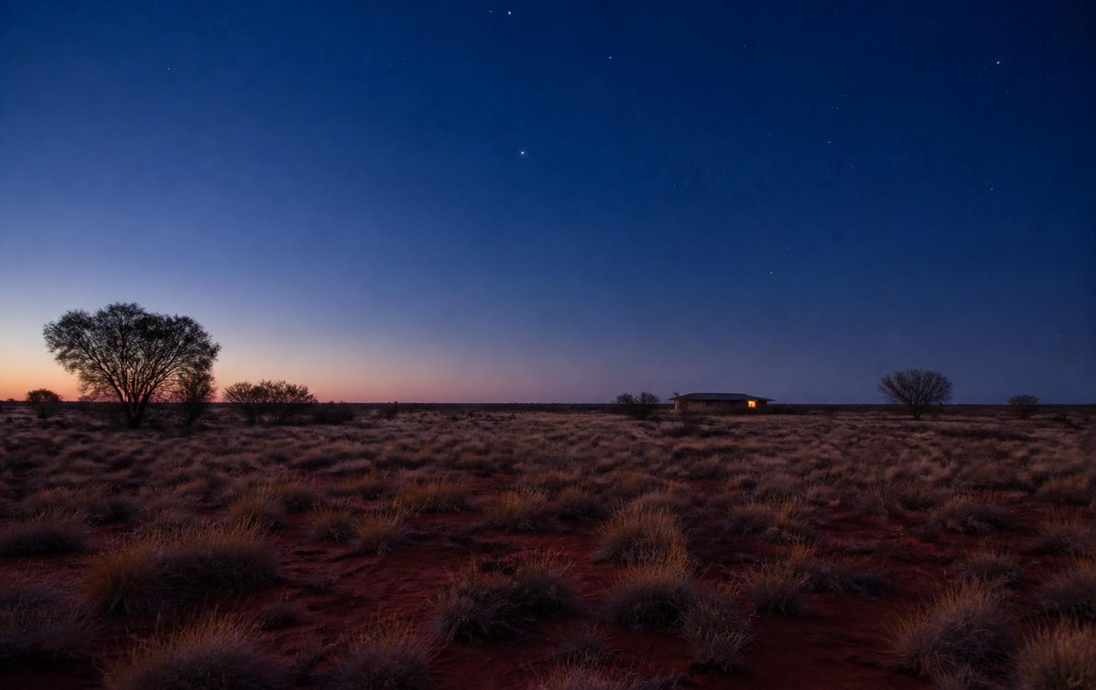 Open red desert plain with spinifex and desert oaks at twilight, a small distant accommodation building showing one warm light