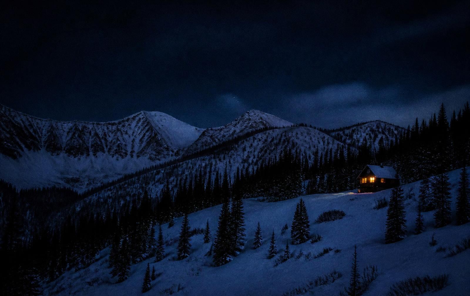 Snow-covered Utah mountain ridges at night with a single warmly lit window among dark pines