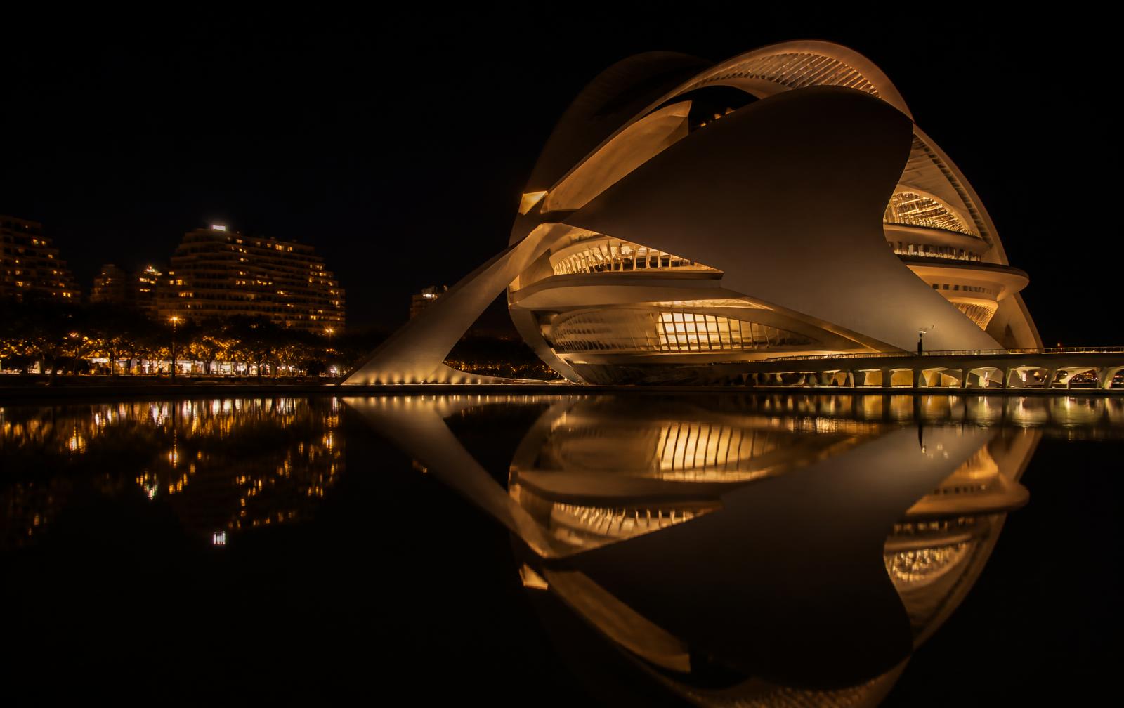 Modern white curved architecture in Valencia lit warm gold at night, reflected in a dark still pool
