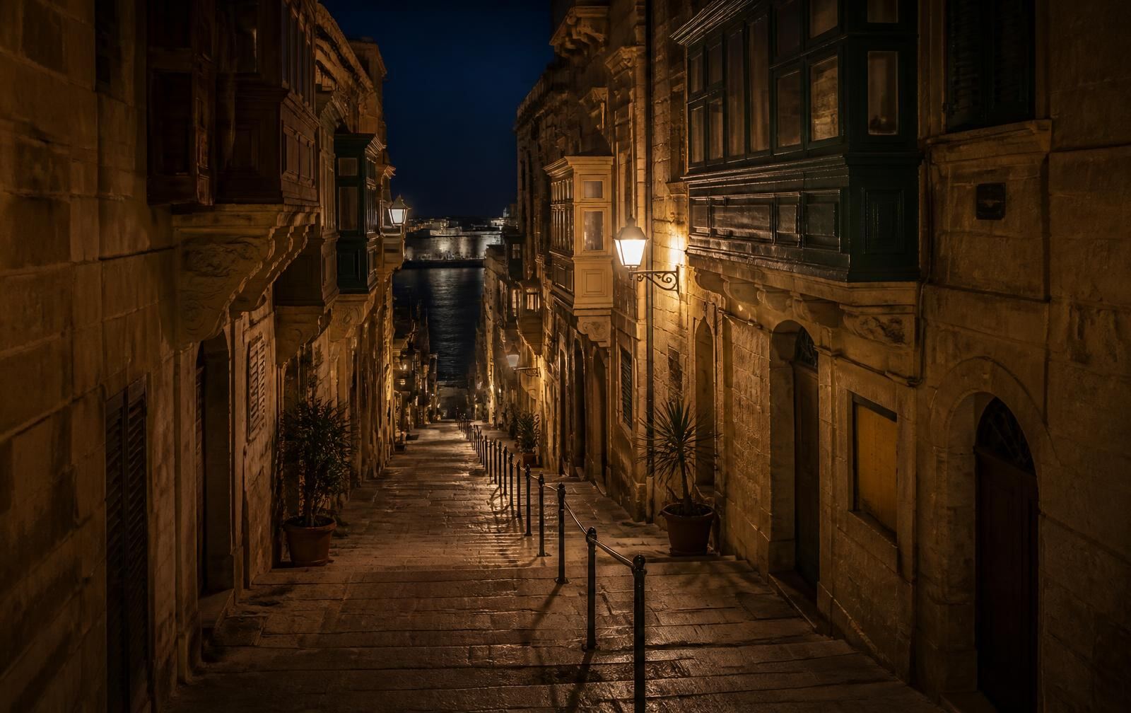 A narrow limestone street in Valletta at night with enclosed wooden balconies, warm lamplight and dark harbour water below