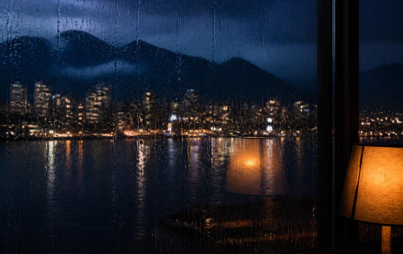 Rain running down a high window in Vancouver at night with blurred waterfront lights and dark mountains beyond