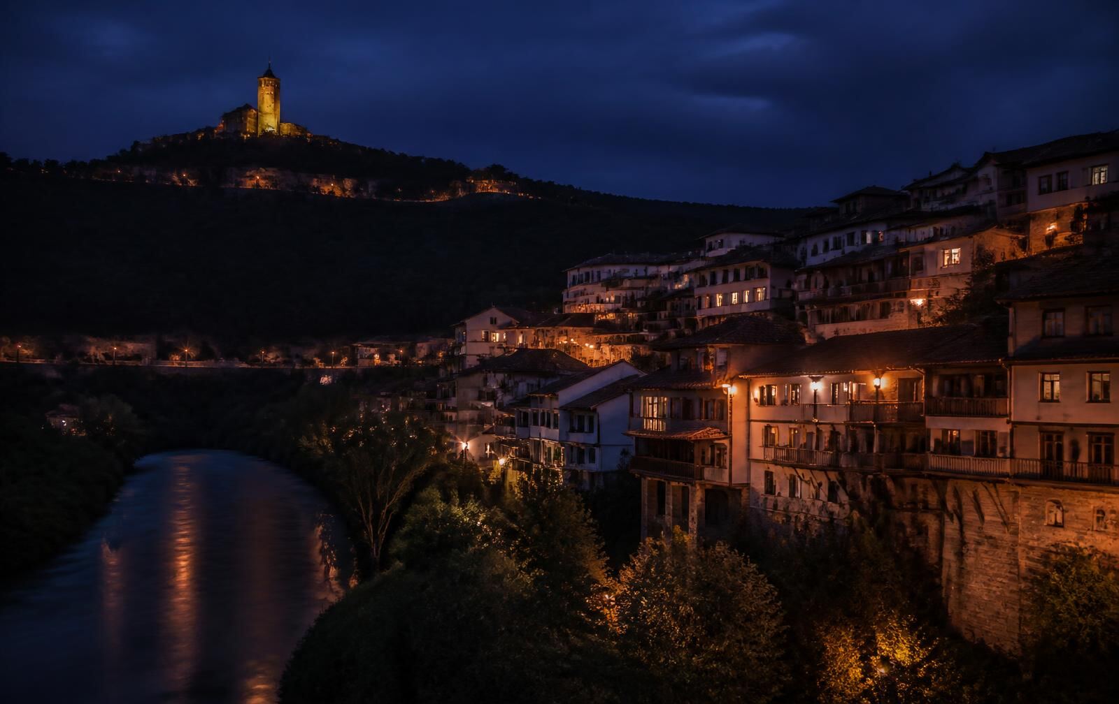 Veliko Tarnovo at night — warm-lit houses stacked above the Yantra gorge with the fortress silhouette on the hill