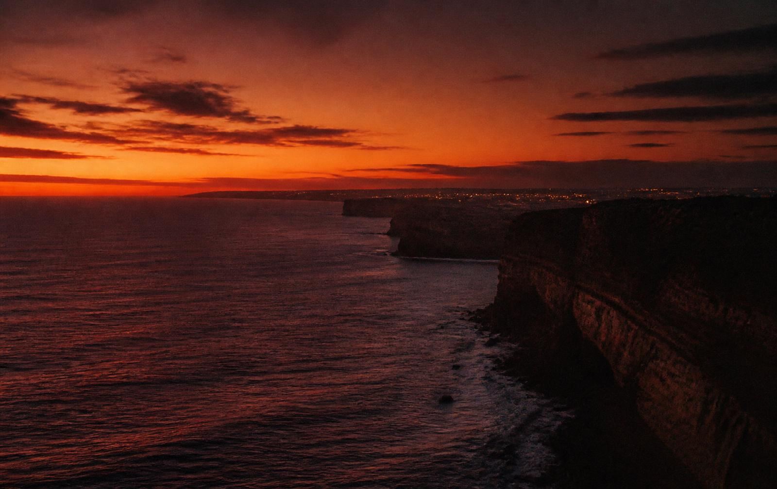 A vast western coastline at deep dusk with burnt-orange horizon, dark cliffs and distant city glow