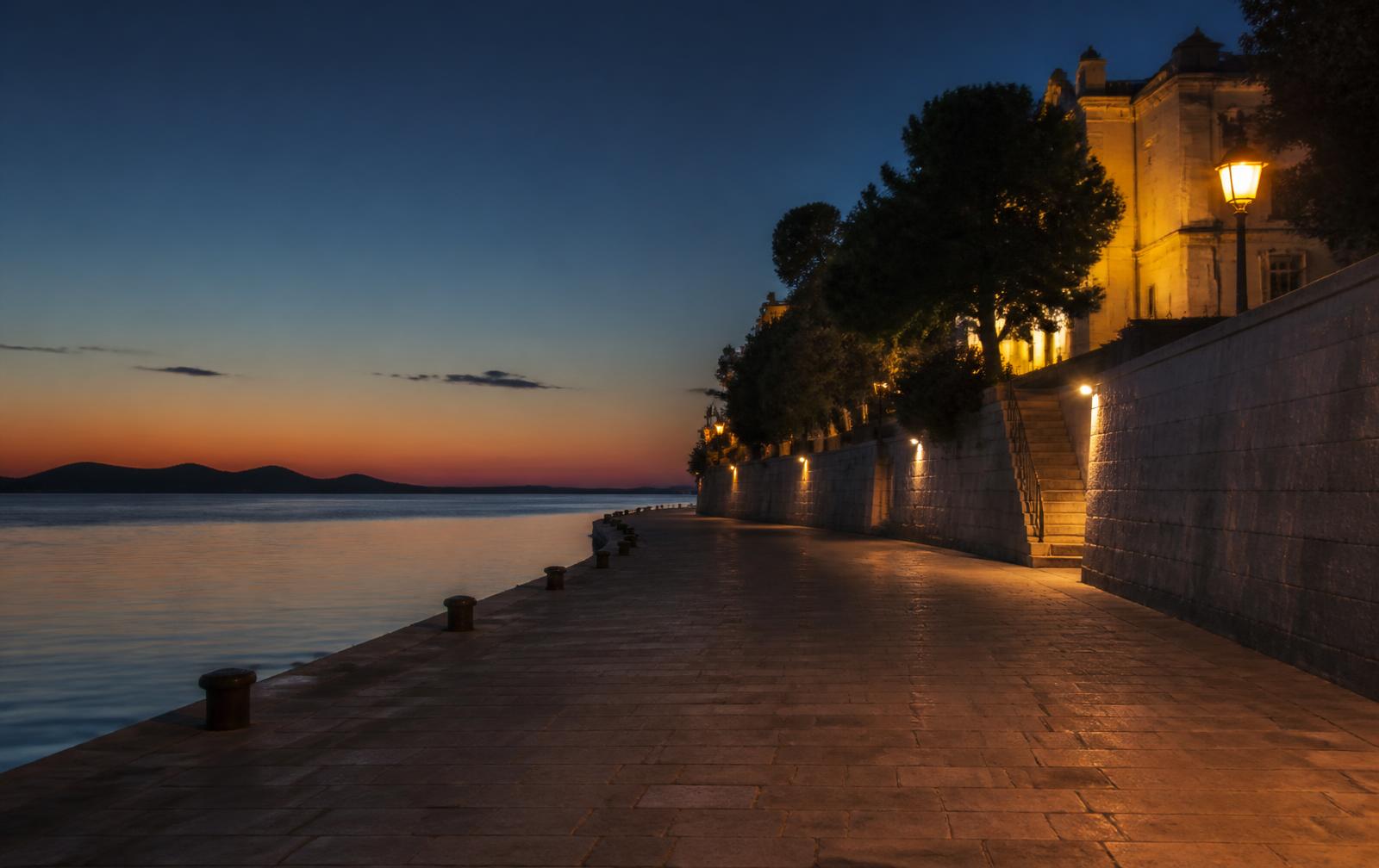 Zadar's waterfront promenade at dusk — warm lantern light on pale stone, dark sea and island silhouettes beyond