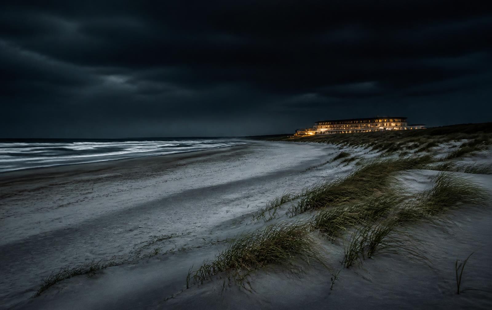 Empty North Sea beach at night with dune grass, dark surf and a distant warmly lit beachfront hotel