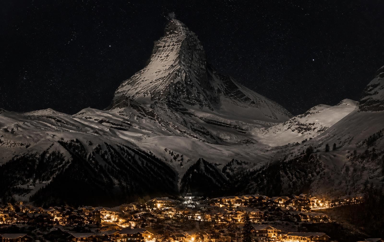 A pyramid-shaped snow peak above a small alpine village lit warm gold under a starry night sky